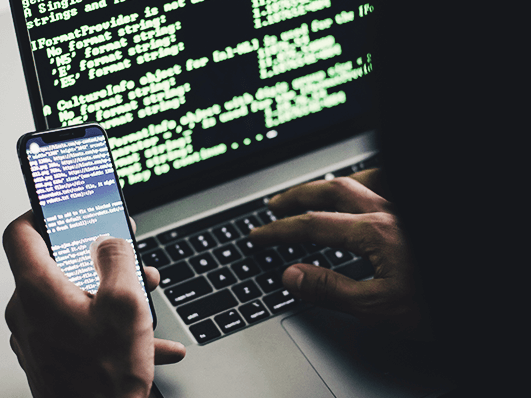 Person am Laptop und mit einem Handy in der Hand beim Code schreiben