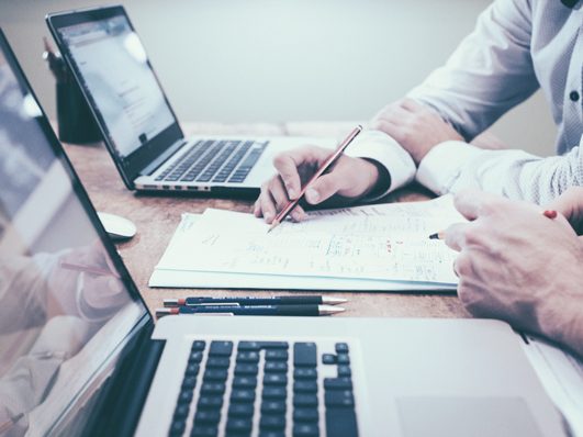 Two people working on laptops and writing on a notepad