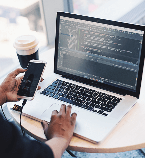 Person mit Handy in der Hand und schreibt auf eine Laptoptastatur Code